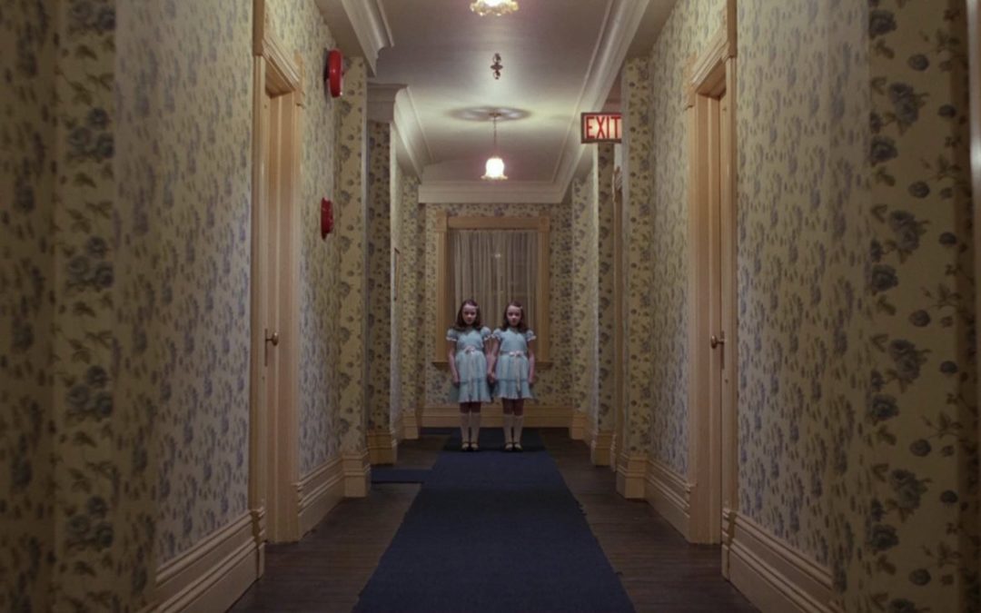 Two twin girls in matching dresses stand at the end of a hotel hallway