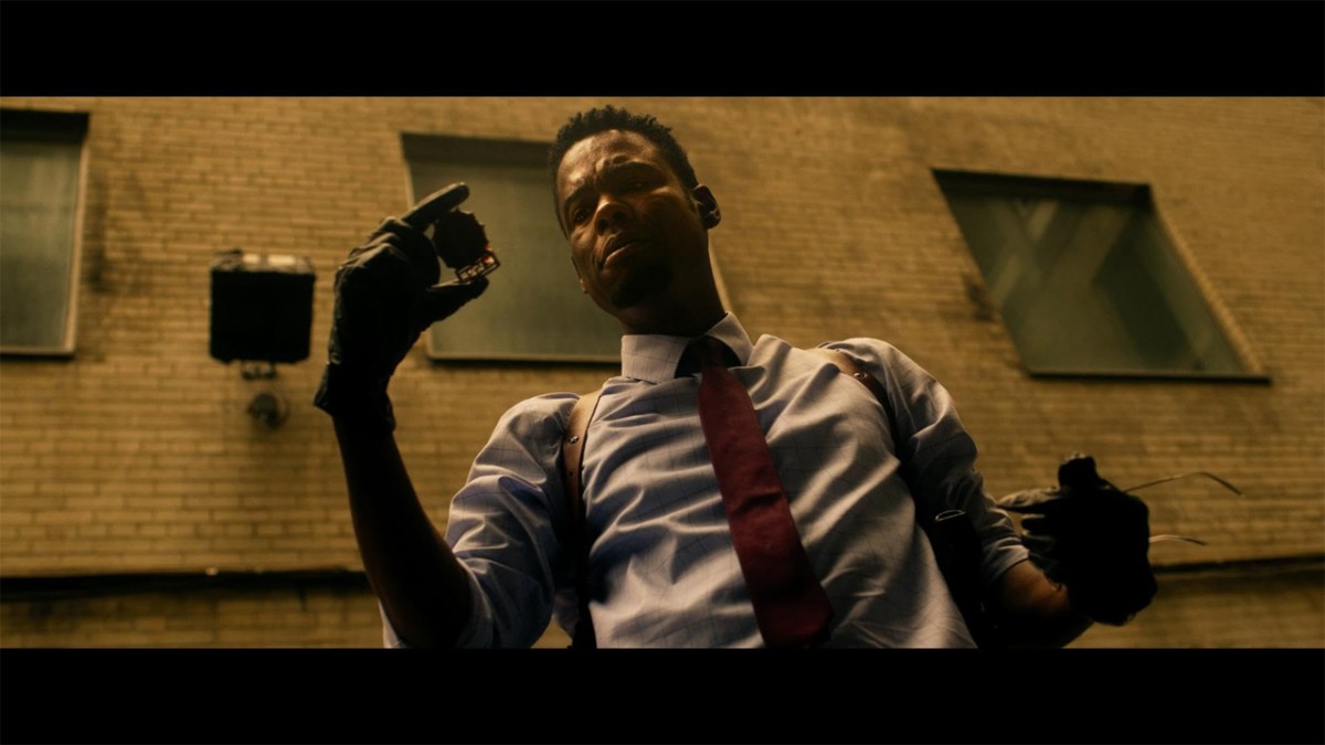 Chris Rock, looking at a strange item in his hand against a sepia-tinted backdrop, taken from the film Spiral.