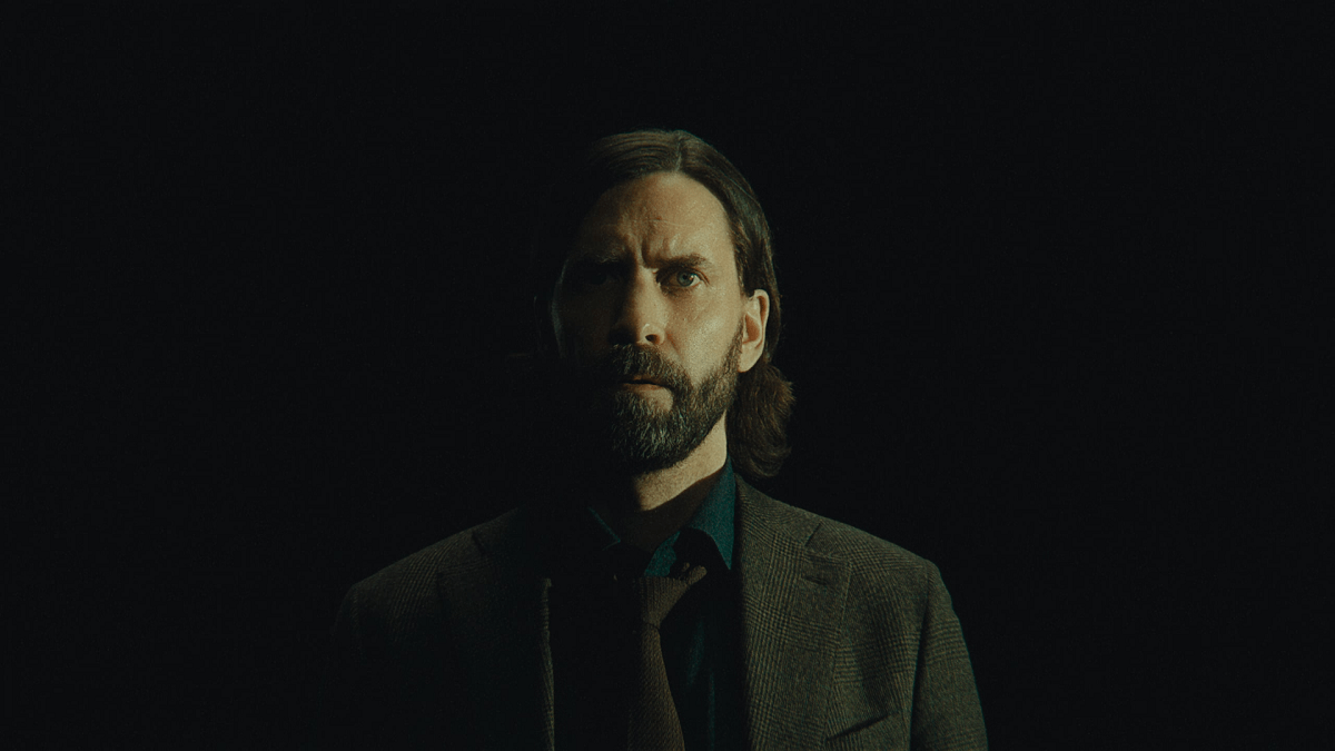 An image of Alan Wake in Alan Wake 2 as part of an article on how Remedy should continue to use the Control style for future games. Alan Wake is standing against a dark background wearing a suit and long, kempt hair. 