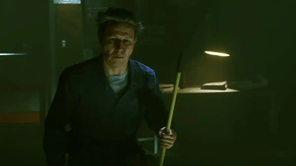 An image of Ahti from Alan Wake 2 as part of an article explaining who the character is in the game and Control. The image shows him standing in a dark room dressed as a janitor and holding a mop. A light is on behind him, illuminating some sort of book.