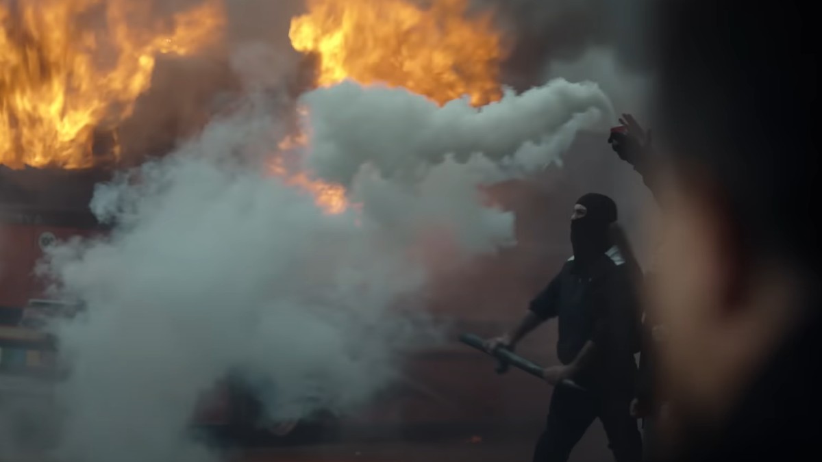 A black-clad rioter against a backdrop of smoke and flame from Suburraeterna.