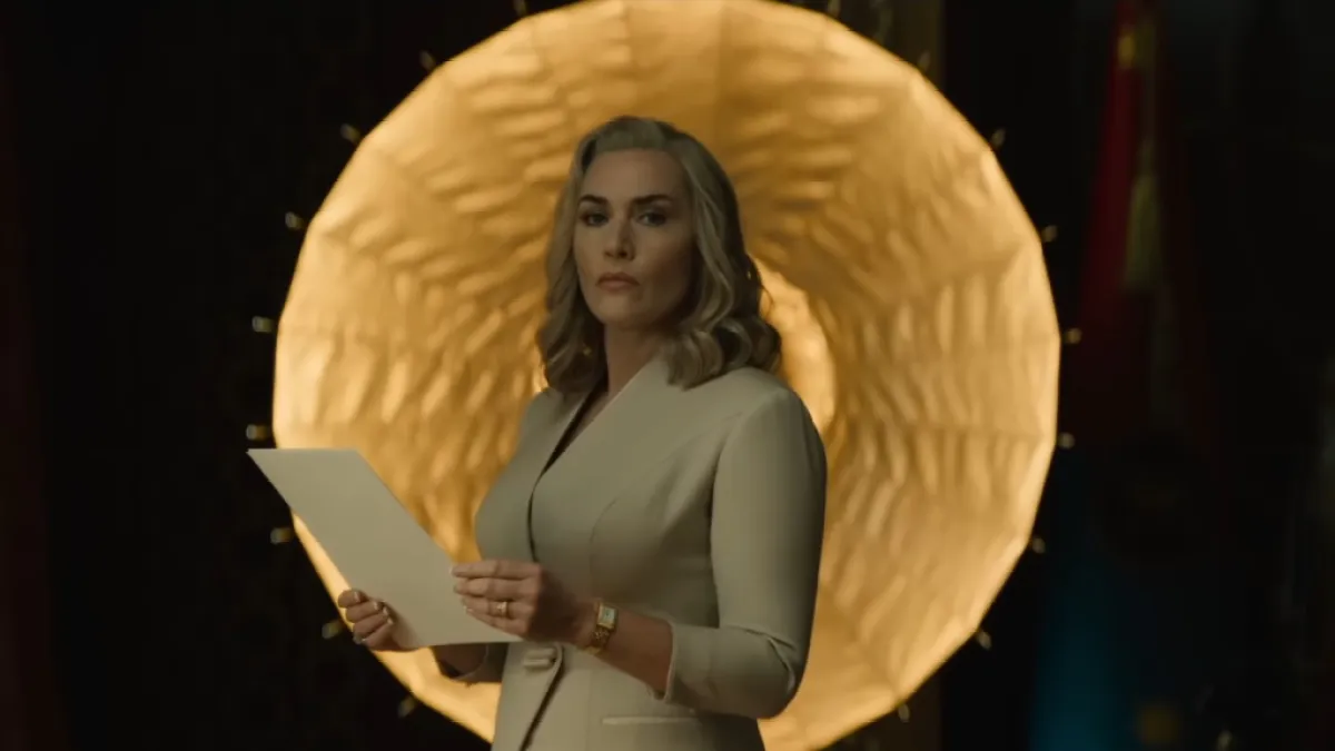 Kate Winslet, as Chancellor Elena Verham, wears a white suit and holds several sheets of paper in a still from The Regime.