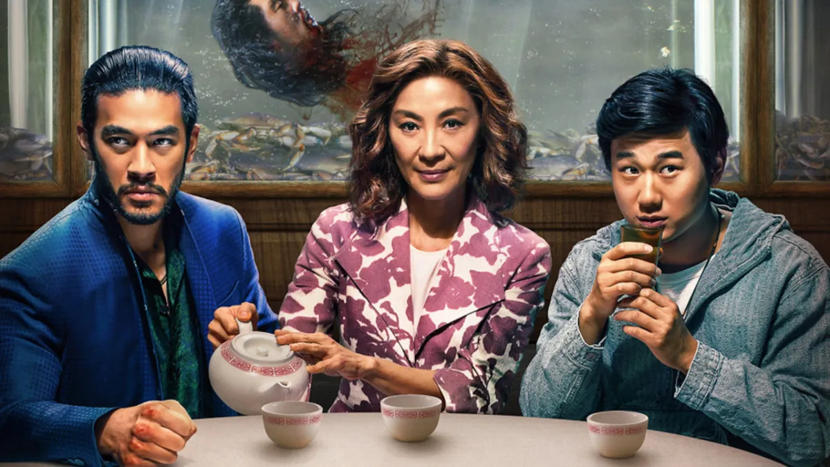 A woman and two younger men sitting at a table, drinking tea. A severed head is floating in a fish tank behind them. This image is part of an article about all the major actors and the cast list for The Brothers Sun.