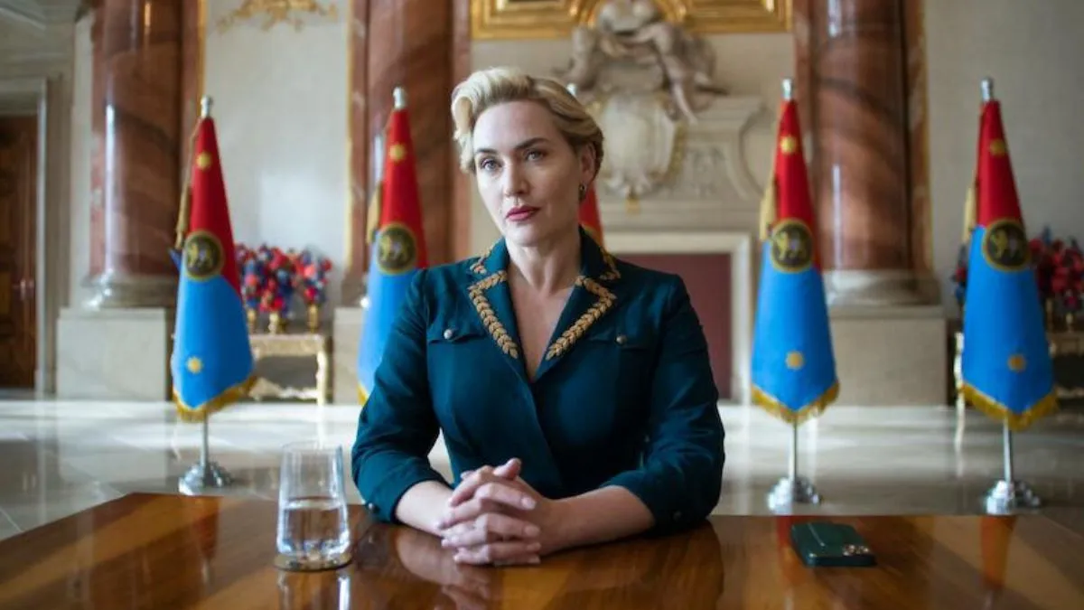 Kate Winslet, as Chancellor Elena Vernham, sits at a table with a series of flags behind her in a still from The Regime.