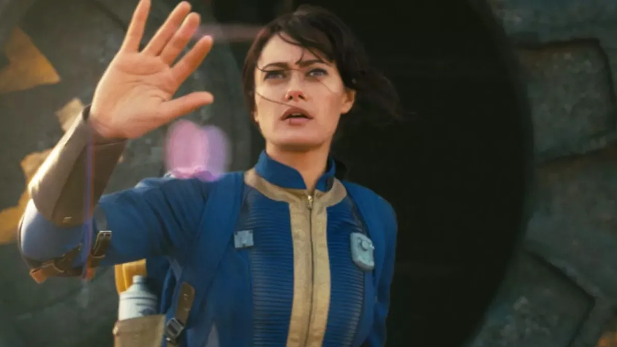 Amazon's Fallout series, a woman in a blue jumpsuit holding her hand up as she looks into the sky.