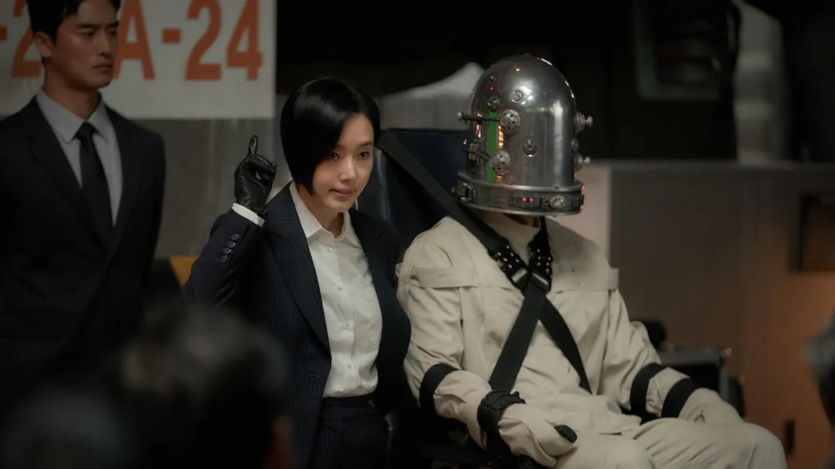 Parasyte: The Grey, a woman in a suit standing next to a humanoid figure in a white prison-style outfit, restrained, with a metal device on their head.