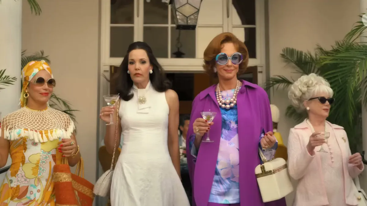 Four women, three of them holding cocktails and wearing sunglasses, exiting a building in 60s/70s clothes. 