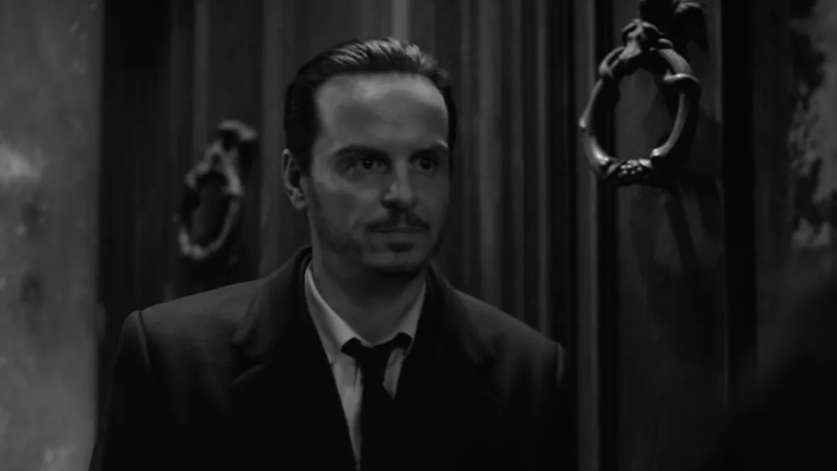 Ripley, played by Andrew Scott, in black and white, looking to the right, standing against a big door.