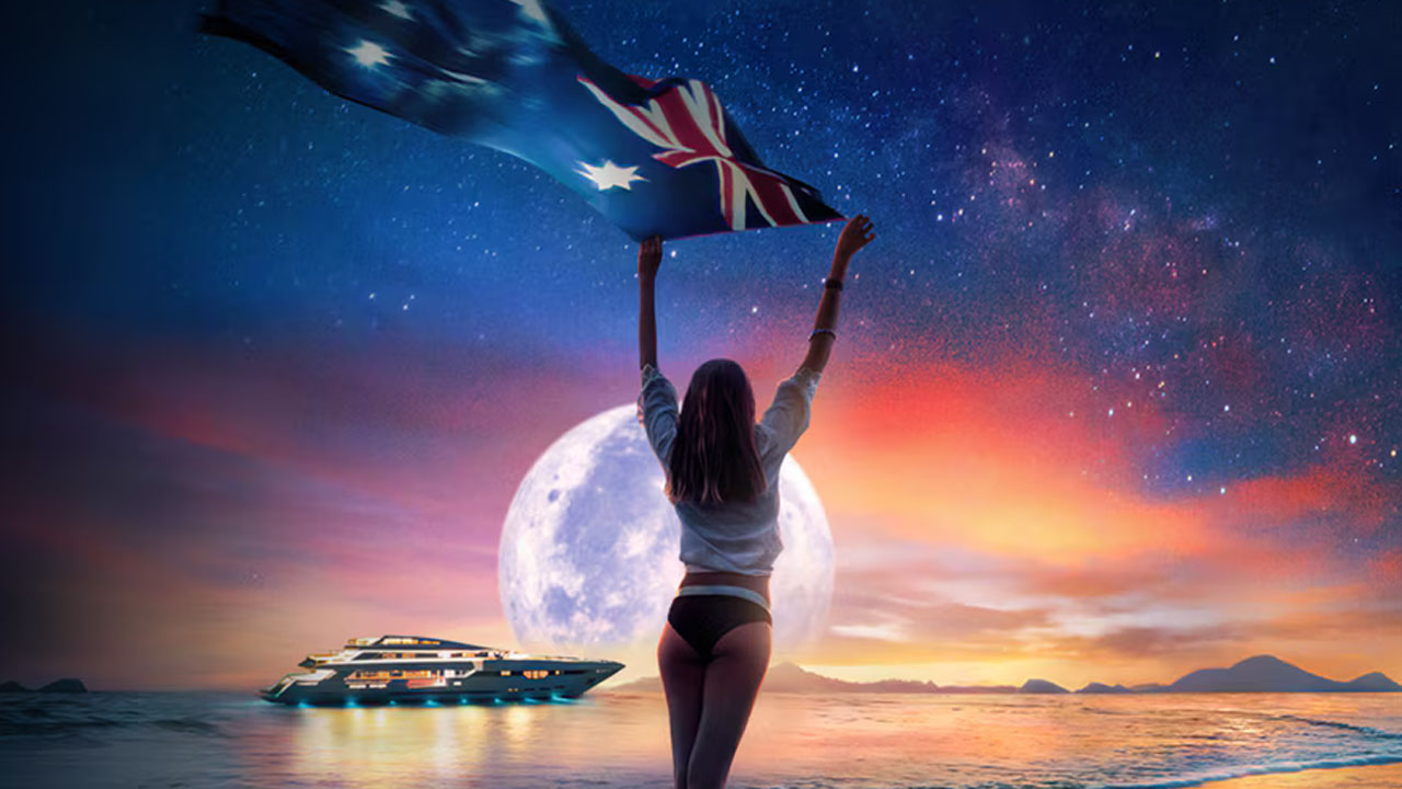 Below Deck Down Under, a woman in a bikini bottom waving an Australian flag on a beach while a yacht goes past in the distance.