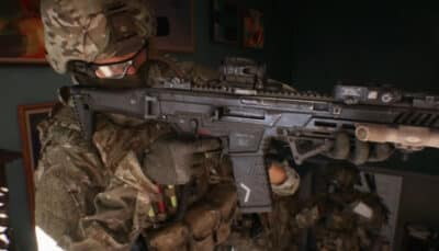 A soldier in camo gear holds a futuristic rifle indoors. The green and brown tones of his uniform blend with dim lighting