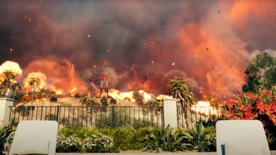 A fiery Battlefield 6 scene showing a massive explosion with smoke and flames rising behind palm trees and a red tower structure.