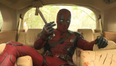 Deadpool in red suit holding knife while sitting in damaged car backseat