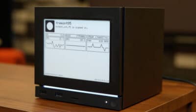 A compact Steam Machine with a built-in E-Ink front display shows system stats like CPU, GPU, and fan speed while sitting on a wooden table