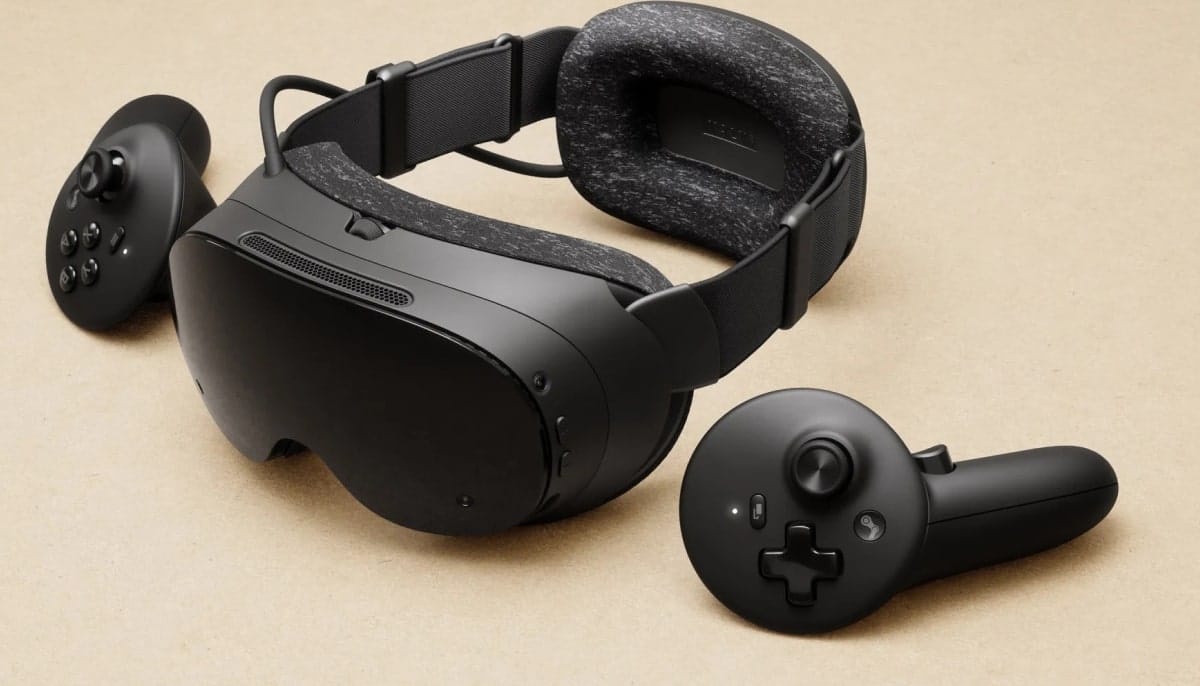 A black Steam Frame VR headset with a padded head strap displayed on a beige surface, accompanied by two matching motion controllers placed on either side