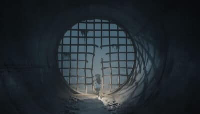Brother and sister protagonists stand inside a dark tunnel facing a broken metal gate on their escape from the island in Reanimal horror game.