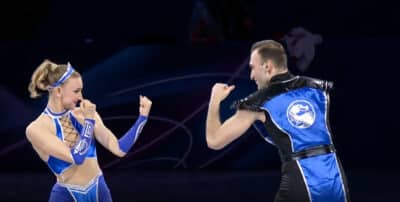 Anastasiia Metelkina and Luka Berulava perform at the Olympic Exhibition Gala in blue Mortal Kombat-inspired costumes, facing each other in fighting stances on the ice.