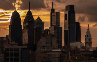 Downtown Philadelphia skyline at sunset with silhouetted skyscrapers against an orange sky, including modern glass towers and the clock tower of City Hall.