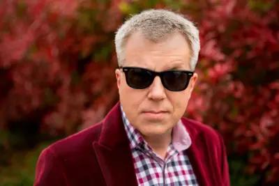 Portrait of Larry Hryb wearing black sunglasses and a red velvet jacket, standing in front of red foliage.